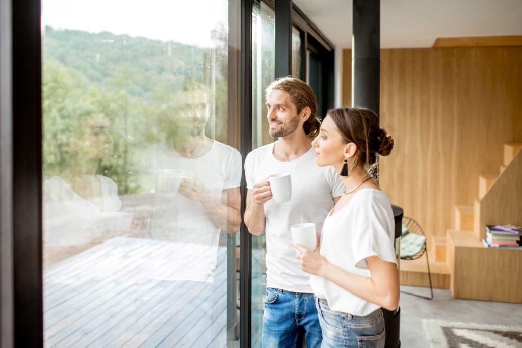 Ein lächelndes Paar steht in einem modernen Zuhause und schaut durch große, klare Fenster nach draußen auf eine grüne Landschaft. Beide halten Tassen in den Händen und genießen die Aussicht.