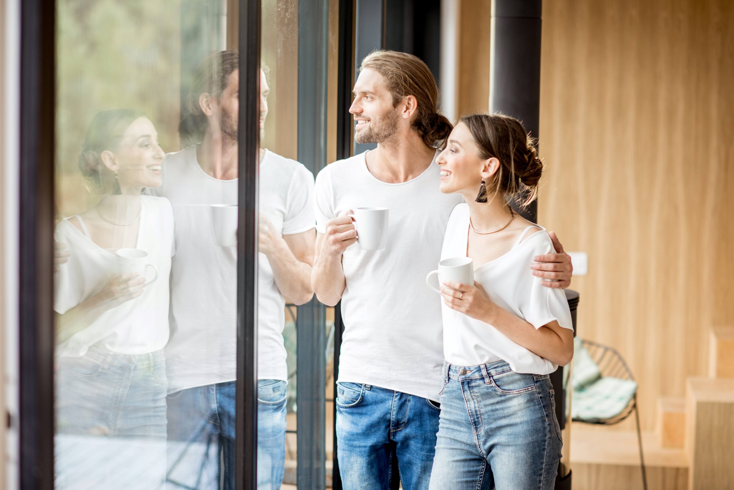 Ein lächelndes Paar steht in einem modernen Zuhause und schaut durch große, klare Fenster nach draußen. Beide halten Tassen in den Händen und genießen die Aussicht, während sie sich umarmen.