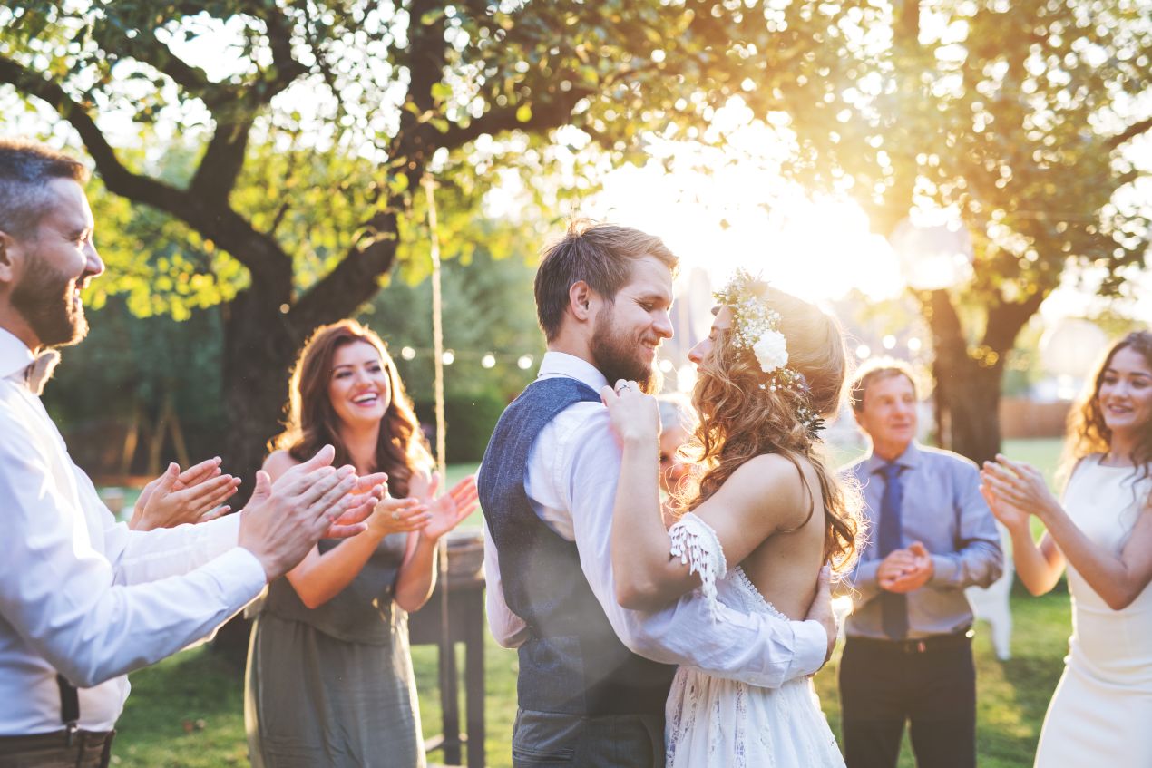 Hochzeit auf dem Bauernhof: Brautpaar tanzt entspannt unter Obstbaeumen im Sonnenlicht, umgeben von applaudierenden Gaesten