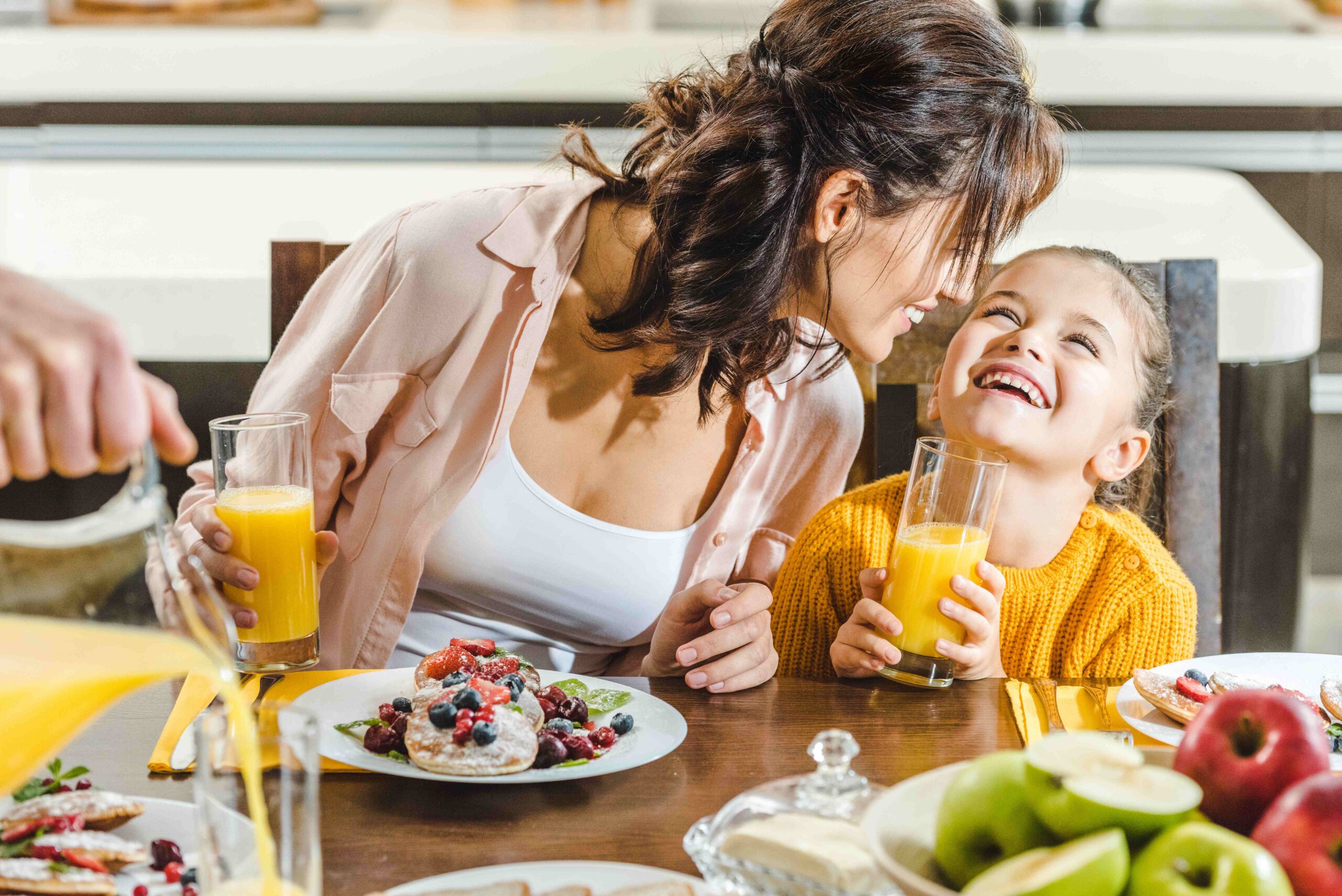 Fröhliche Mutter mit Tochter sitzt am Tisch mit frischem Saft und Pfannkuchen mit Beeren in der Küche